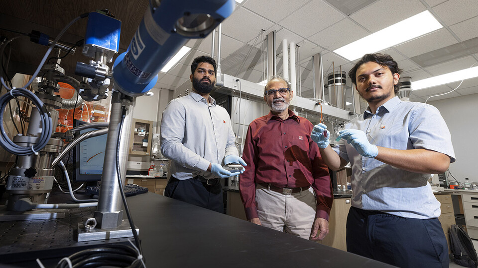 Abhijeet Prasad (left), a doctoral student in engineering; Ravi Saraf (center), Anderson Distinguished Professor of chemical and biomolecular engineering; and Aashish Subedi, a senior physics major, are photographed in Saraf’s Othmer Hall lab. Prasad is holding small electronic chips used to study the nanoparticle necklace network devices. Subedi is holding a suspension of nanoparticle necklaces (blue container) made from individual particles (red container). (Craig Chandler / University Communication)