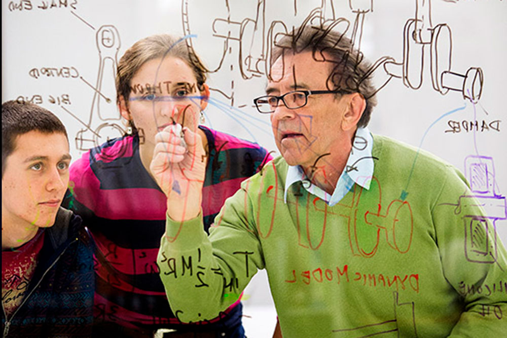 Wieslaw Szydlowski explains the design of an engine to students Matt Thompson (left) and Maggie Clay as it is drawn on the glass walls of the Olsson Room inside the Engineering Library in Nebraska Hall. (University Communication photo)