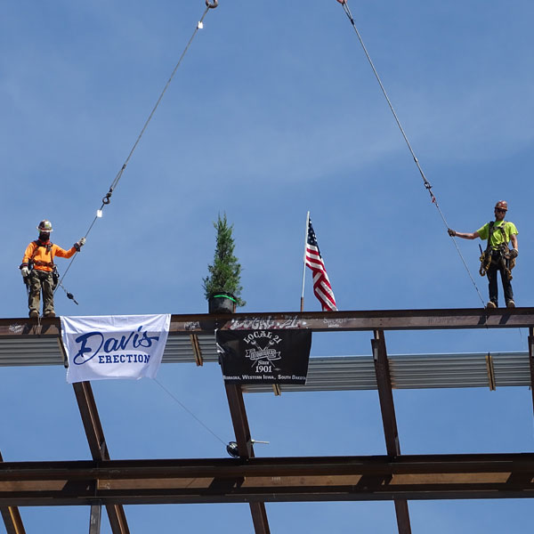 The final beam is secured on top of Kiewit Hall, with 2 workers and tree posing standing on top (Aug 12, 2022).
