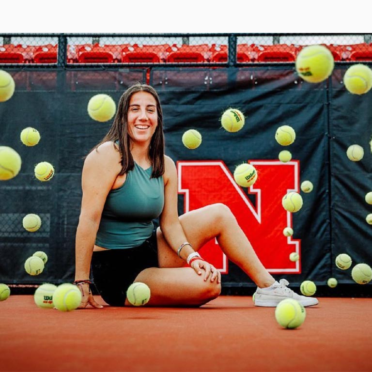 Isabel Adrover Gallego, May 2023 computer engineering graduate and Husker tennis player sitting on the court with tennis balls all around her.