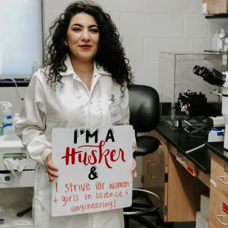 Woman in lab coat holding a sign that says: I'm a Husker & I strive for women and girls in science and engineering.