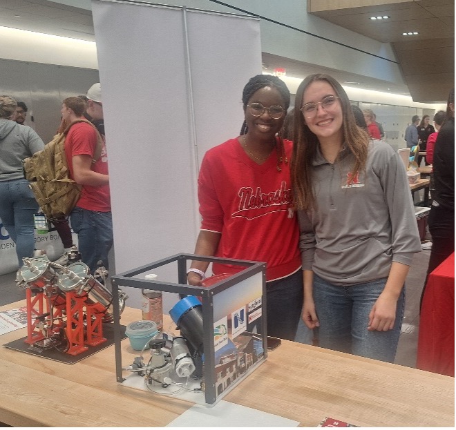 Two people standing behind a table displaying engineering models at an indoor event.