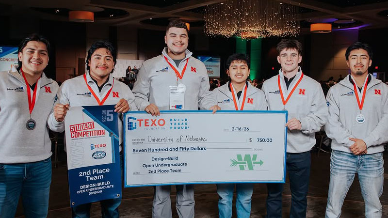 Group of people in matching gray sweaters holding an oversized check and medals in an indoor setting.