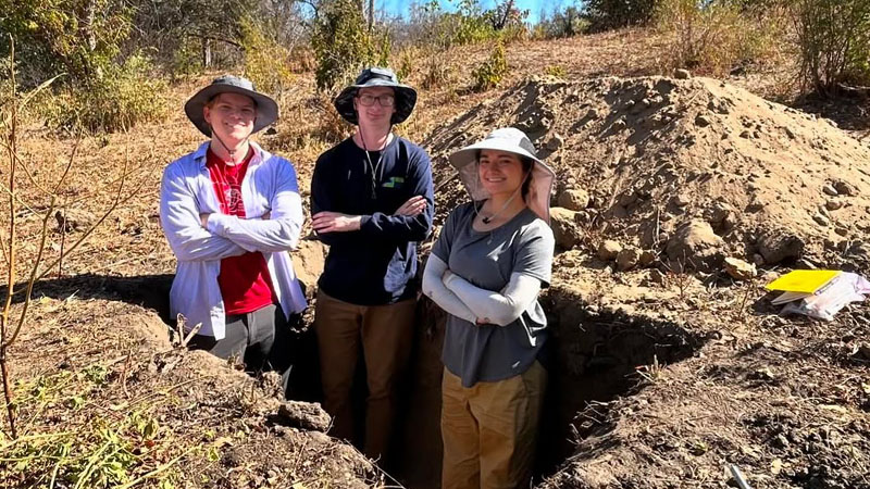 Group of people standing in a dug-out trench in a dry, outdoor setting with surrounding vegetation.