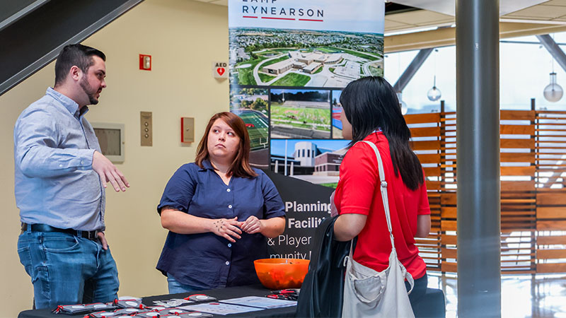 Group interacting at an event table with informational materials and banner in background.