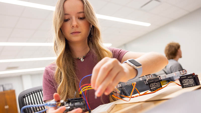 Person assembling wires in a classroom setting with electronic components on a table.