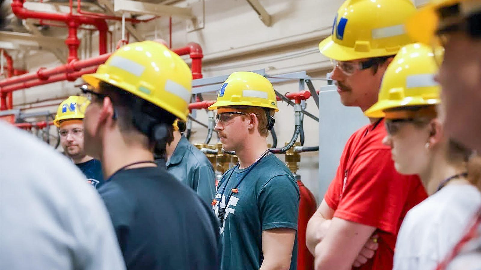 Students wearing yellow hard hats gathered in an industrial setting near red pipes.