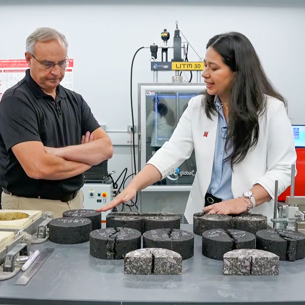 Two people examining cracked cylindrical asphalt samples on a table in a laboratory setting.