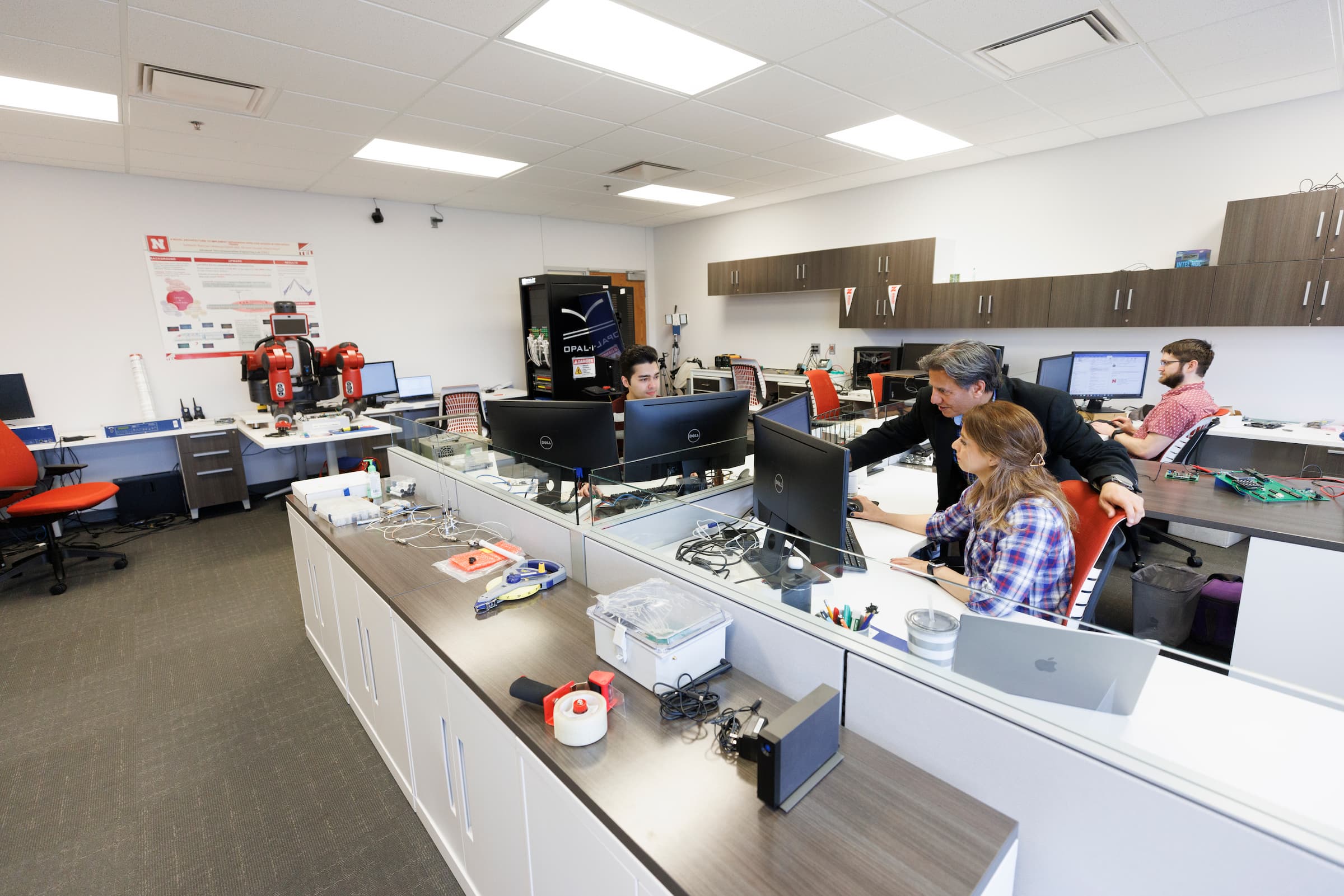 Office space with people working at desks, computers, and equipment on countertops.
