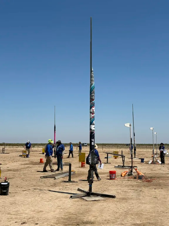 A tall rocket on a launch pad in a dry field, with people and equipment in the background.