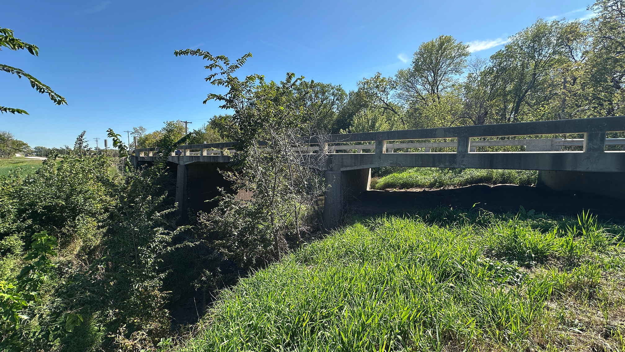 Concrete bridge over a grassy ditch surrounded by trees under a bright blue sky.
