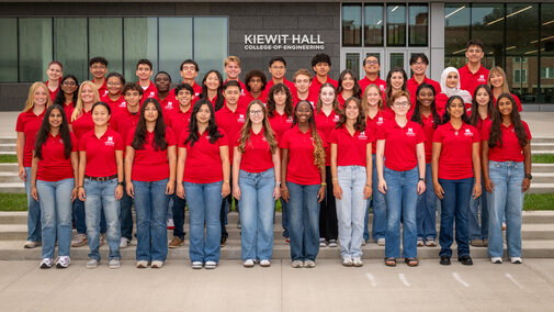 Forty University of Nebraska–Lincoln students in red polos and blue jeans pose for a group photo in front of Kiewit Hall.
