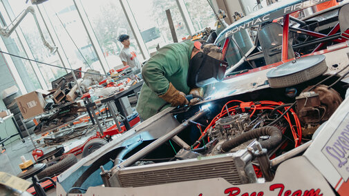 Mechanic welding a race car in a workshop, surrounded by tools and equipment.