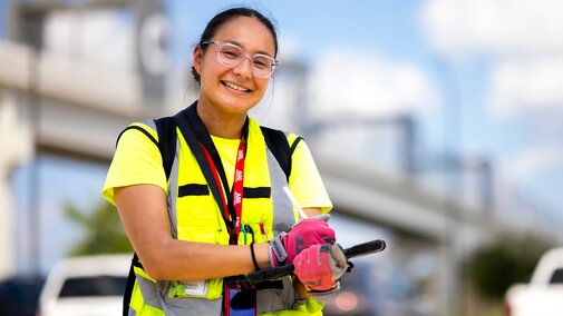 Smiling worker in a yellow safety vest and gloves, holding tools, with a blurred urban background.