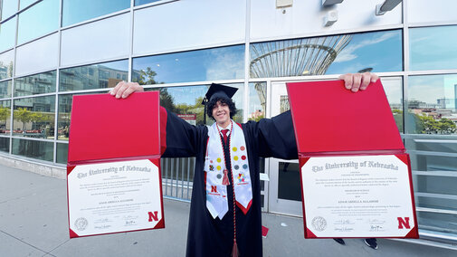 Graduate holding two diplomas outside a building, wearing a cap and gown with bright sashes.