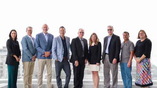 Group of nine people standing outdoors on a rooftop, dressed in business and casual attire.