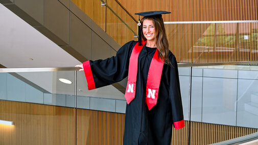 Graduate in black robe and cap with red accents stands in a modern building with glass and wood decor.