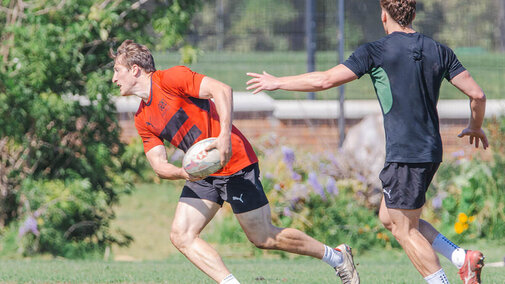 Two men playing rugby on a grassy field; one holding the ball, evading the other's defense.