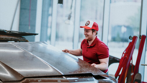 Man in red shirt and cap inspecting a car hood in a brightly lit workshop.