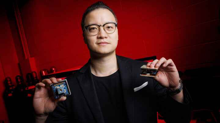 Jun Wang, assistant professor of electrical and computer engineering at the University of Nebraska–Lincoln, holds two variants of the novel 10 kilovolt silicon carbide MetaPak switch cells inside his lab in the Scott Engineering Center. He is wearing a dark blue sport coat over a black shirt and glasses. The lab is lit with red light.