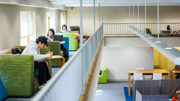 Bright indoor study space with students working on laptops and seated on green chairs.