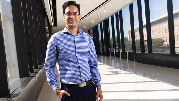 Man in a blue checkered shirt standing in a sunlit hallway with large windows.