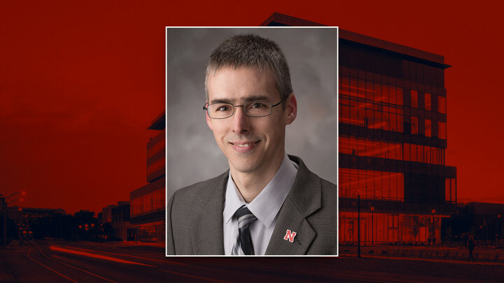 Man in a suit and tie with an "N" pin, red tinted background showing a modern building at dusk.