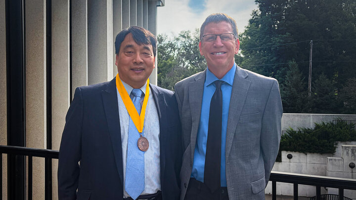 Two men in suits smiling outdoors, one wearing a medal with a yellow ribbon.