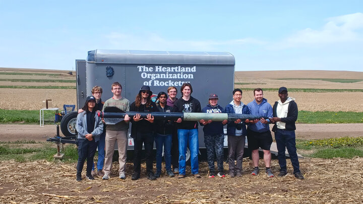 A group poses outdoors holding a rocket, with a trailer and farmland in the background.