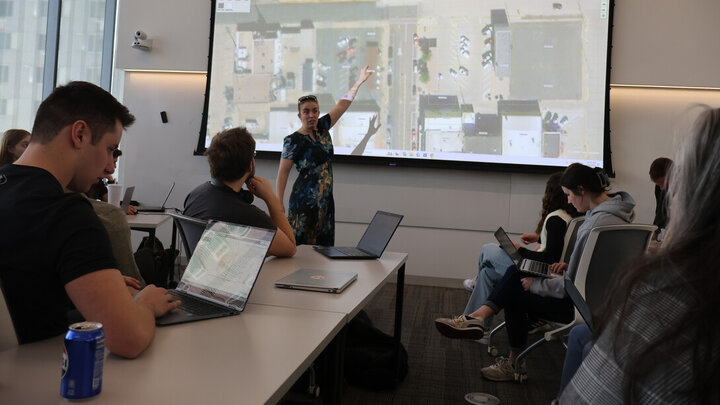 Classroom setting with instructor pointing at a projected map, students seated with laptops.