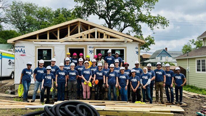 Group of volunteers in blue shirts and helmets standing in front of a partially built house.