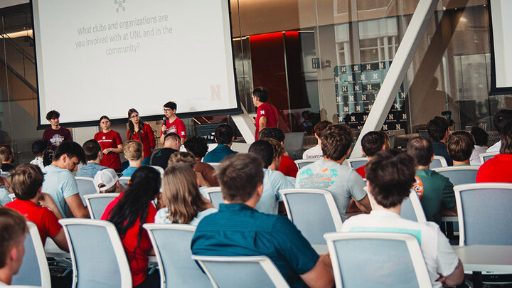 Students sit in a classroom while a group of people present in front of them with a large whiteboard.