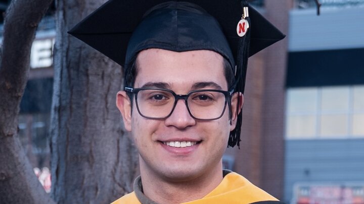 Graduate in cap and gown smiling outdoors near a tree, with a building in the background.