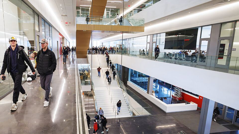 Modern indoor atrium with stairs, glass walls, and people walking and seated at tables.