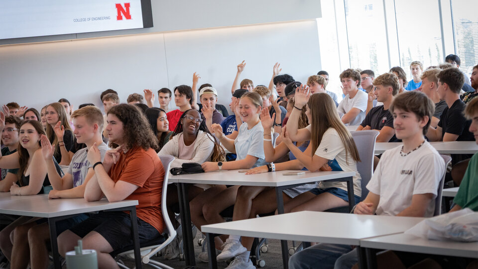 Students seated in a classroom, some raising hands, with bright natural lighting.