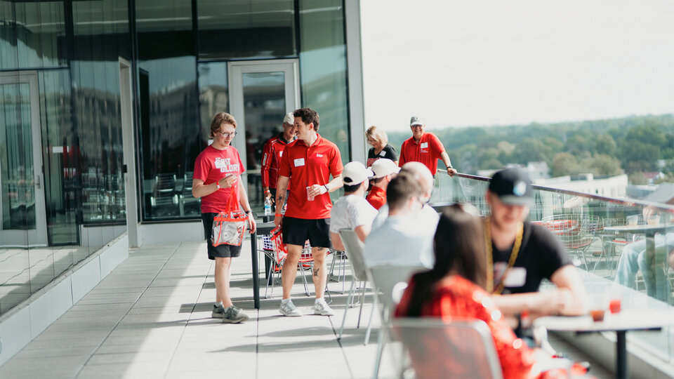 People in red and white converse on a sunny rooftop terrace with glass walls and distant greenery.