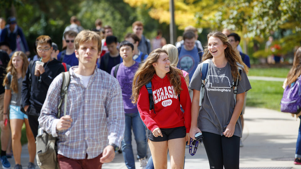 Students walking outdoors on campus, smiling and chatting amidst a crowd, colorful trees in background.