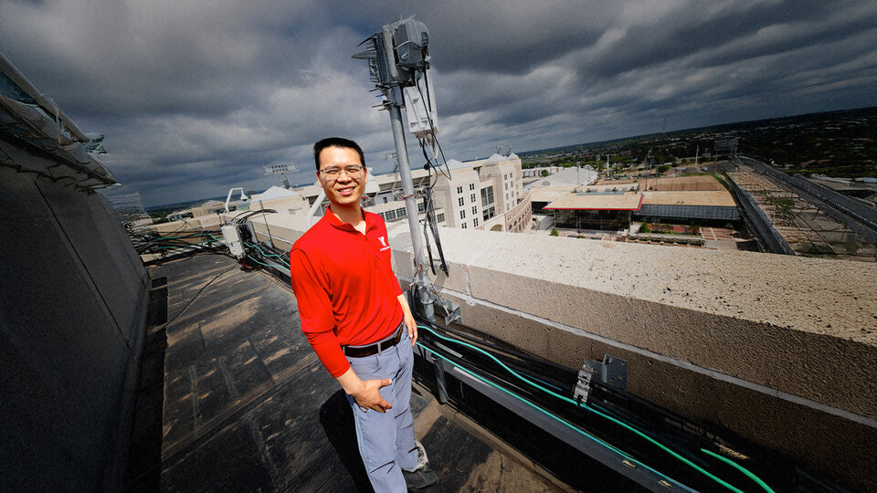 Man in a red shirt standing on a rooftop near telecom equipment, with a cityscape in the background.