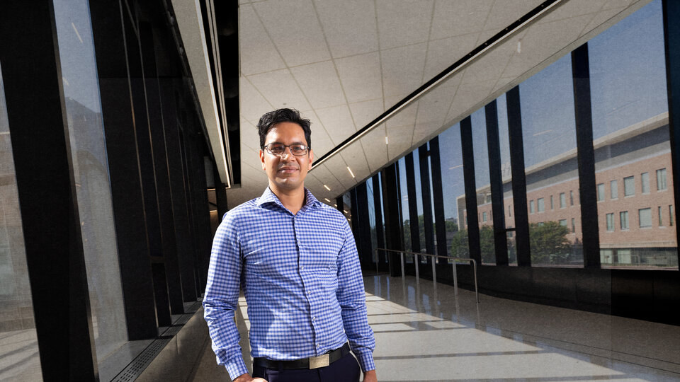 Man in a blue checkered shirt standing in a bright hallway with large windows and a reflective floor.