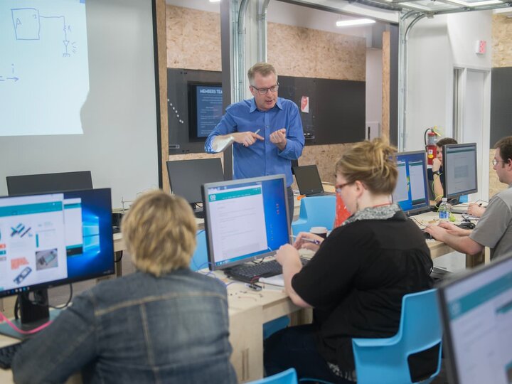 Instructor stands at the front of a classroom, gesturing towards students seated in rows working at desktop computers.