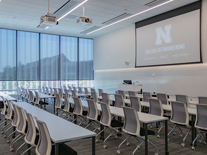 Modern classroom with rows of white desks, gray chairs, large windows, and a projected screen.