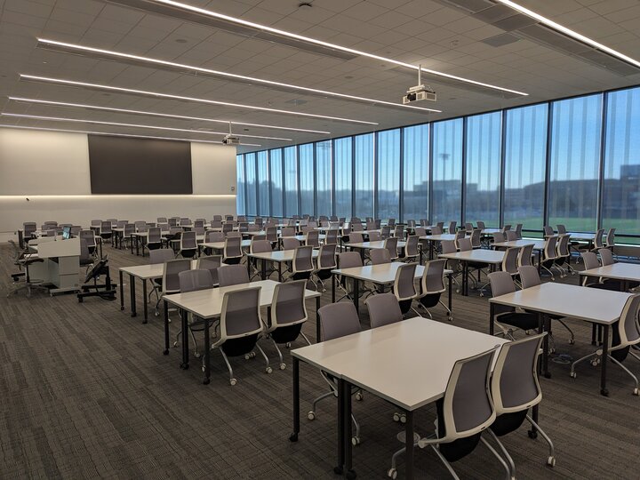 Spacious classroom with rows of tables and chairs, large windows, and a projector ceiling mount.