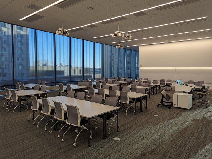 A modern classroom with rows of tables, chairs, ceiling projectors, and large blue-tinted windows.