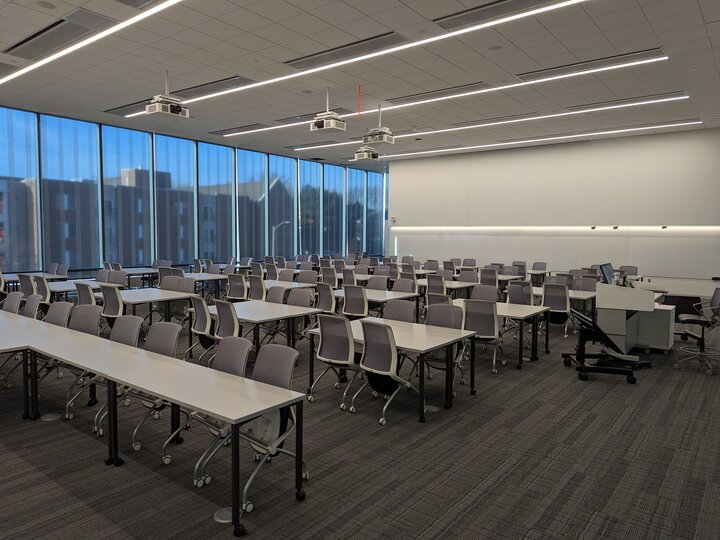Large classroom with rows of tables, chairs, ceiling projectors, and a wall of blue-tinted windows.