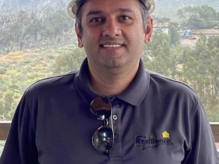 Man wearing a white hard hat and gray polo shirt, standing outdoors with trees in the background.