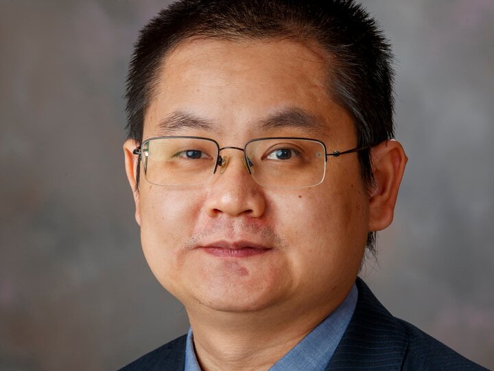 Man in glasses wearing a suit, with a neutral expression and gray background.