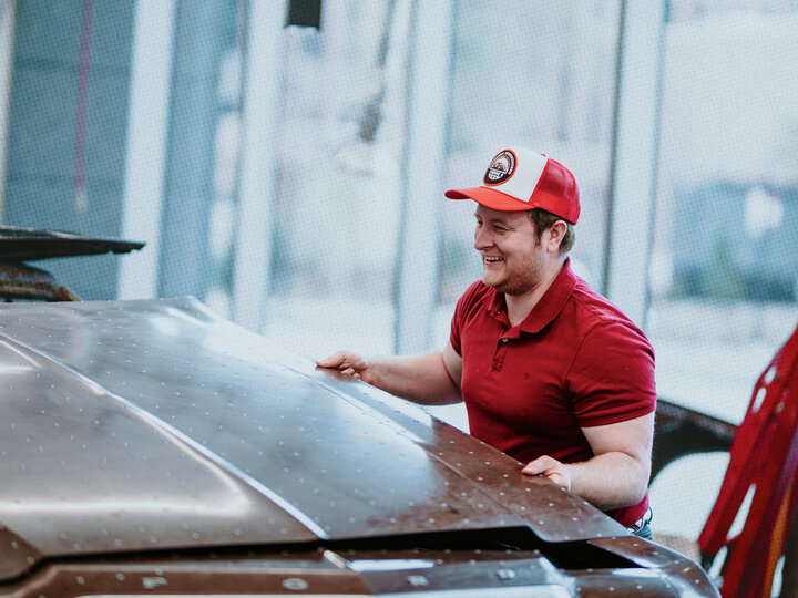 Man in red shirt and cap inspecting a car hood in a brightly lit workshop.