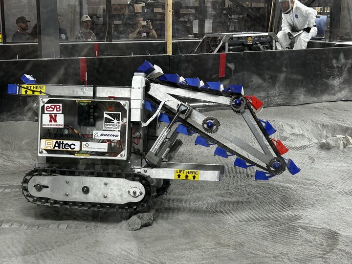 A small robotic vehicle digging gray sand in an enclosed area, with people in the background.