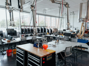 Bright laboratory workspace with tables, industrial equipment, 3D printers, and large windows.