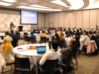 Audience seated at tables in a conference room, a speaker presenting with a projector screen.